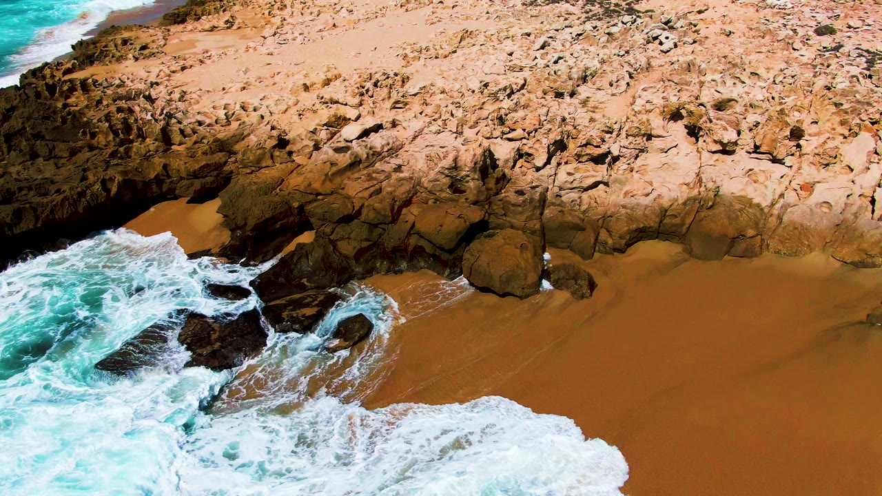 At Punta Buzzards in Baja California Sur, a stronger band of white foam pushes over the lower edge of the beach while dark rocks hold the center of the frame, with no visible people.