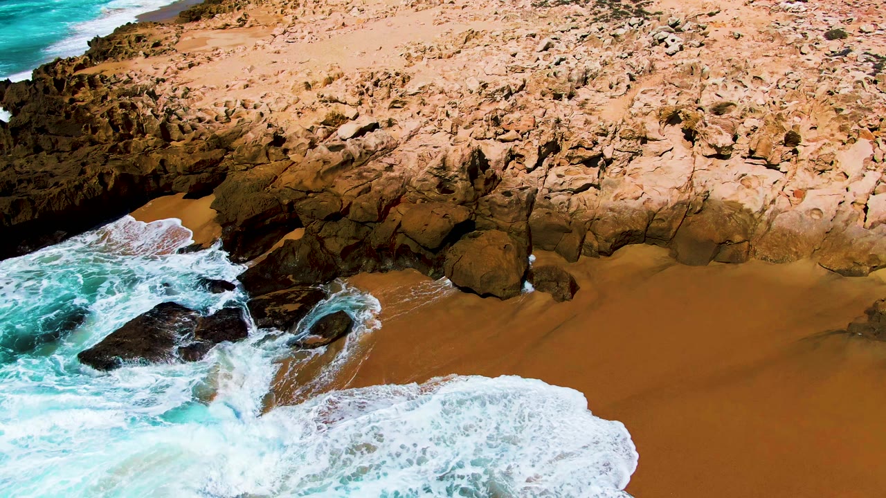 At Punta Buzzards in Baja California Sur, broad whitewater covers more of the lower sand while turquoise water churns around black rocks near the left side, with no visible people.