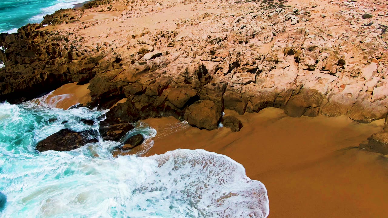 At Punta Buzzards in Baja California Sur, foamy water thins around exposed dark rocks and leaves wet orange sand visible below the rugged tan rock face, with no visible people.