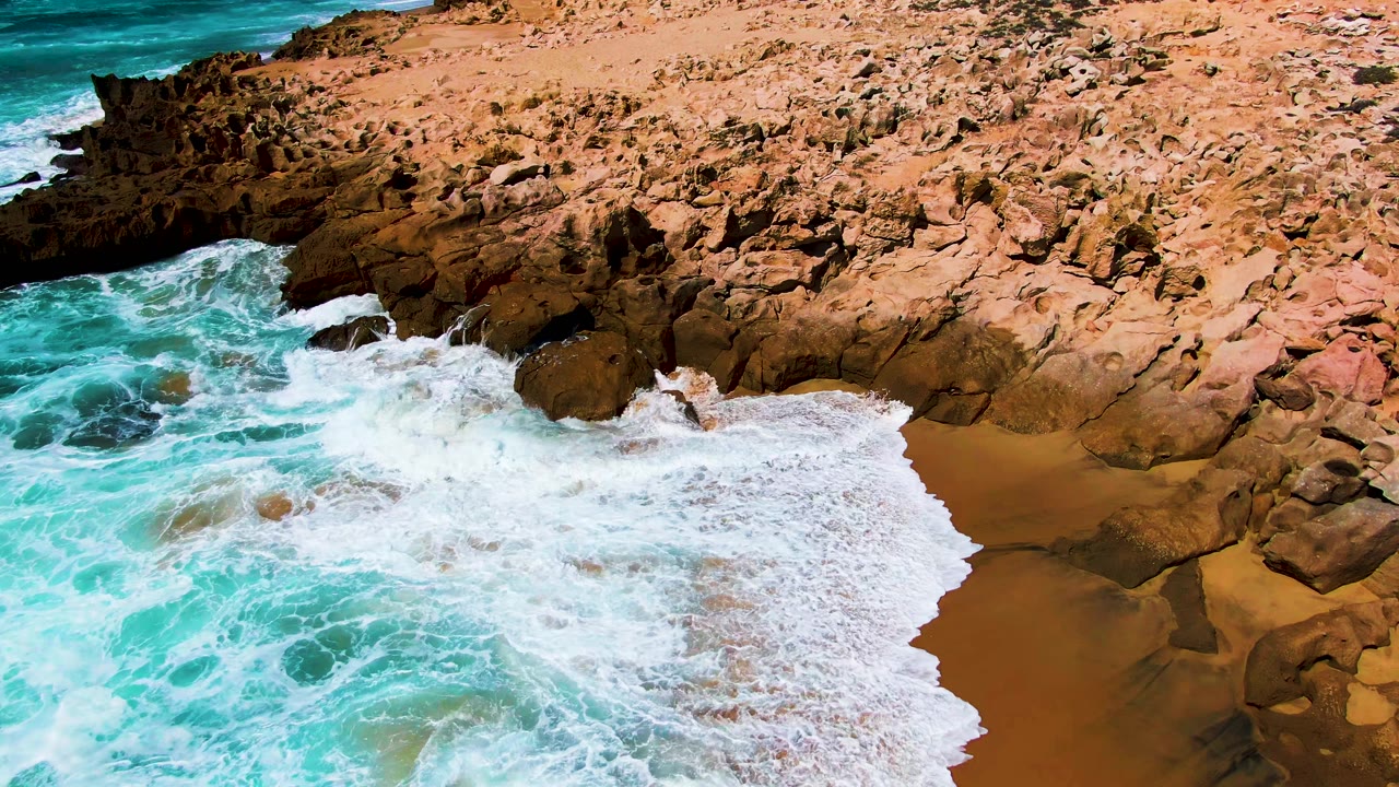 Aerial view of turquoise surf spreading white foam into a rocky cove at Punta Buzzards, Baja California Sur, with tan rock above and no visible people.