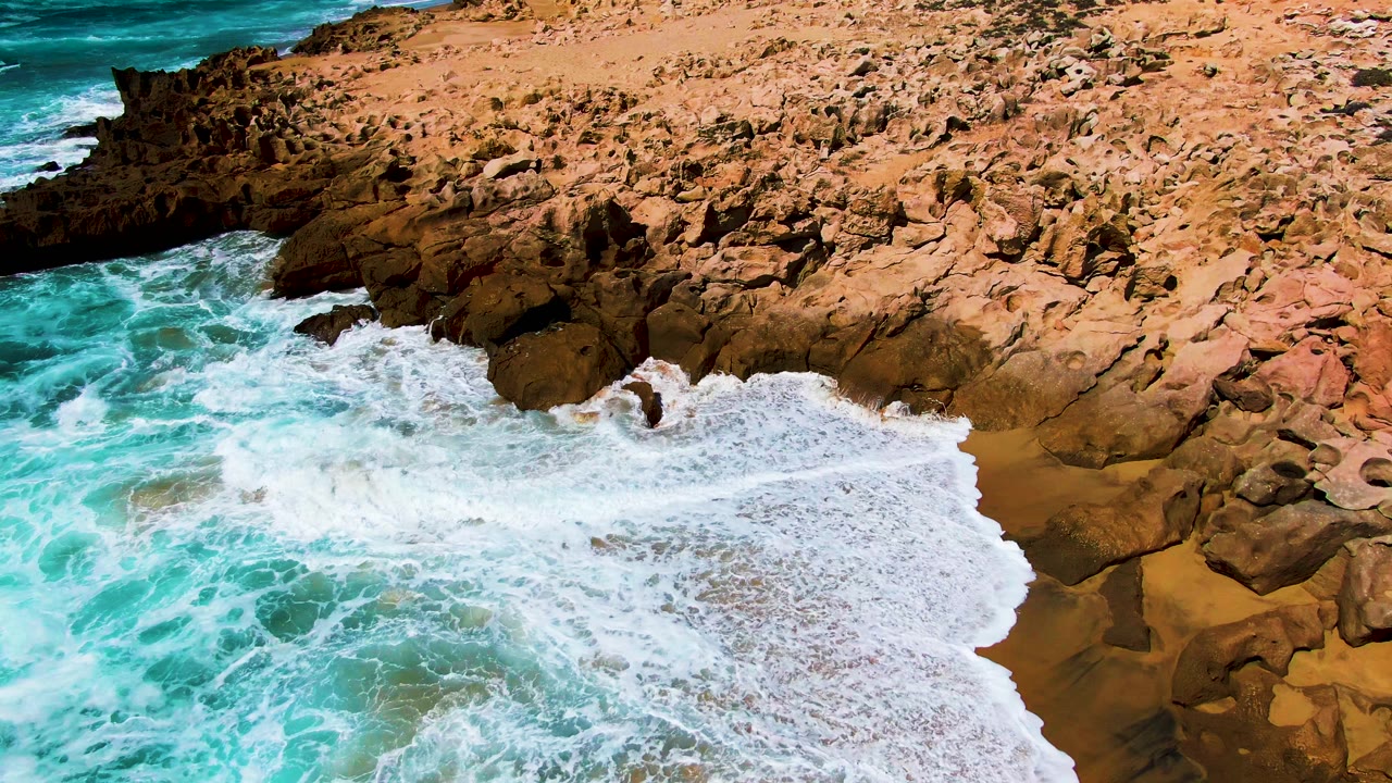 White ocean foam pushes farther into the Punta Buzzards rocky shoreline, covering more of the wet sand while turquoise water fills the lower frame and no visible people appear.