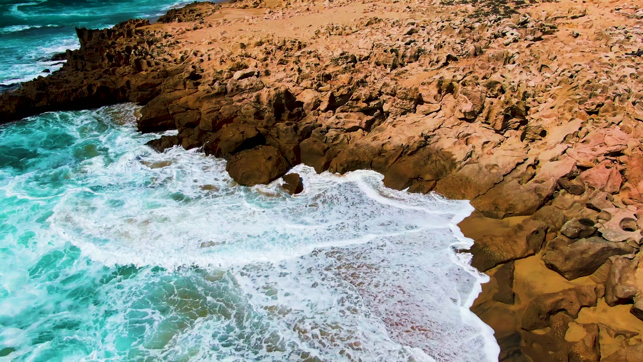 A broad sheet of foamy surf reaches across the rocky Punta Buzzards cove, with dry tan stone at the top of frame and saturated blue-green water at the left, no visible people.