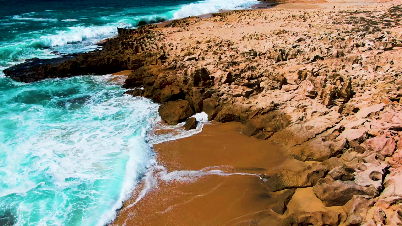 At Punta Buzzards in Baja California Sur, turquoise surf washes around dark rocks and a sandy pocket beach, with no visible people.