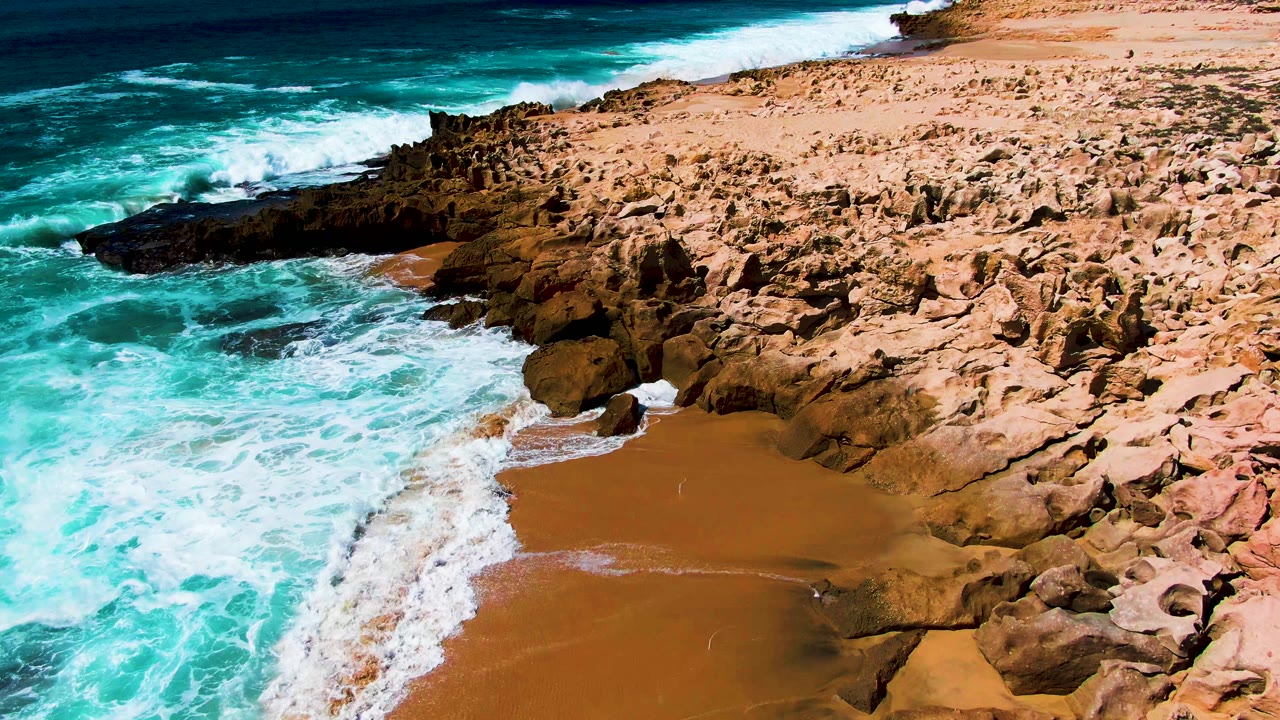 At Punta Buzzards in Baja California Sur, white foam spreads farther into the cove while rough brown rocks frame the sandy beach, with no visible people.