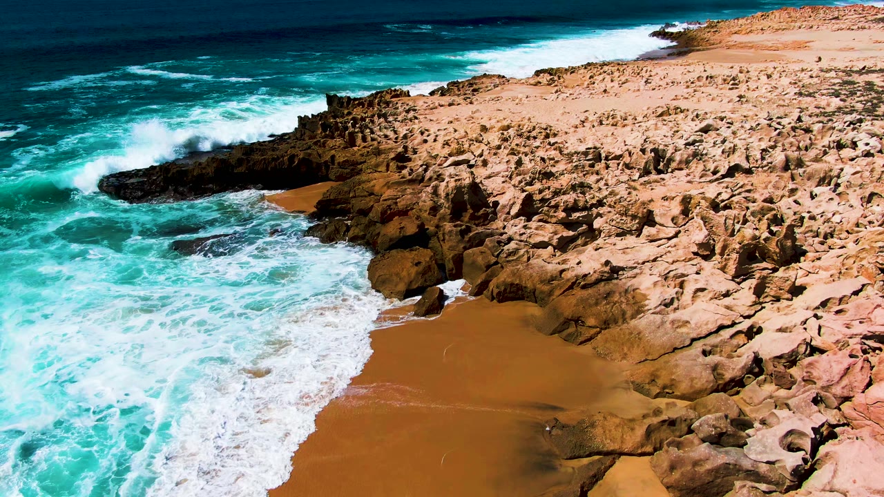 At Punta Buzzards in Baja California Sur, a broad sheet of white surf reaches the sand below the rocky point, with deep blue ocean beyond and no visible people.