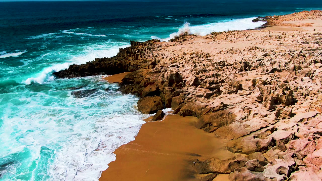 Aerial coastal view with breaking surf, sand, and jagged brown rocks.