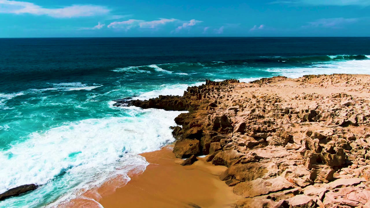 At Punta Buzzards in Baja California Sur, turquoise surf breaks beside tan rock formations and a small sandy beach with no visible people.
