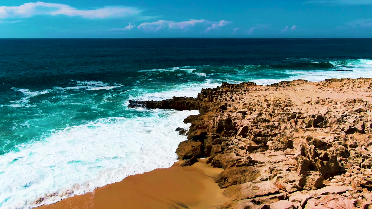 At Punta Buzzards in Baja California Sur, foamy water covers more of the lower beach beside rugged tan shoreline rocks, with no visible people.