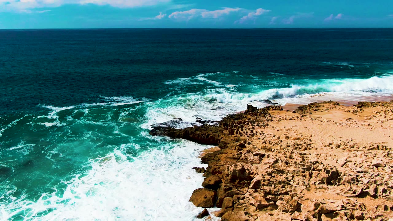 Drone aerial view of the flat rocky coastal shelf at Punta Buzzards, Baja California Sur, Mexico, with turquoise water and white wave foam along the shore edge filling the frame; no sky visible, no visible people
