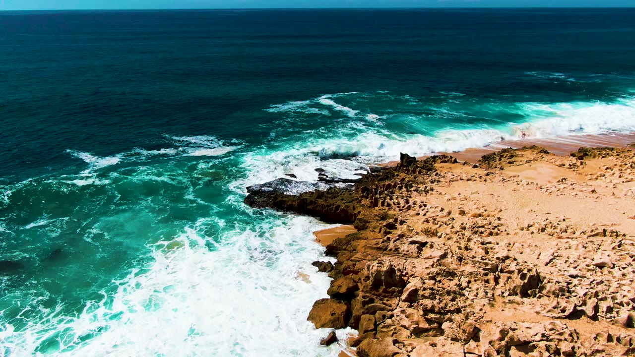 Aerial view from a steep angle above the rocky coastal shelf at Punta Buzzards, Baja California Sur, Mexico, showing turquoise ocean and white surf along the shore edge; no visible people