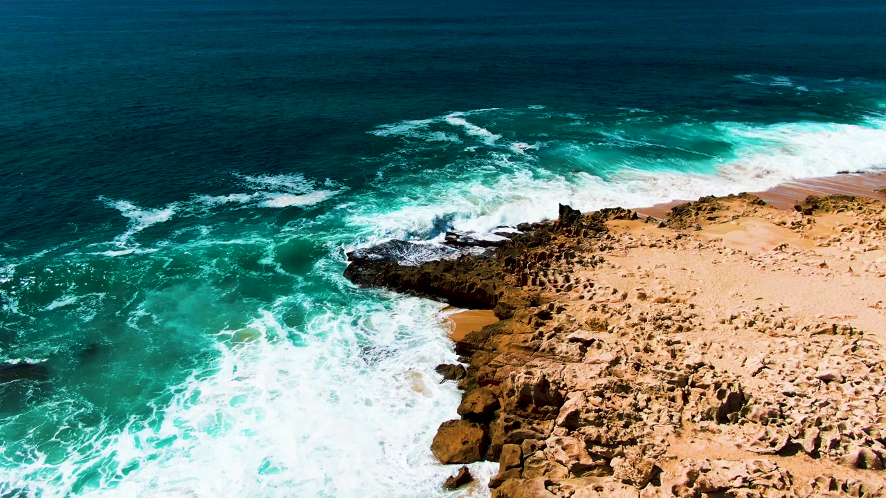 Drone aerial view of the rocky coastline edge at Punta Buzzards, Baja California Sur, Mexico, with turquoise ocean waves actively breaking against the flat rocky shore; no visible people