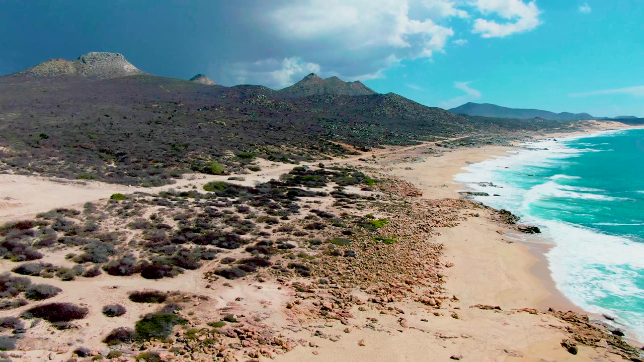 Opening aerial frame over Punta Buzzards in Baja California Sur, with rocky sand and scrub in the foreground, turquoise surf on the right, mountains in back, and no visible people.