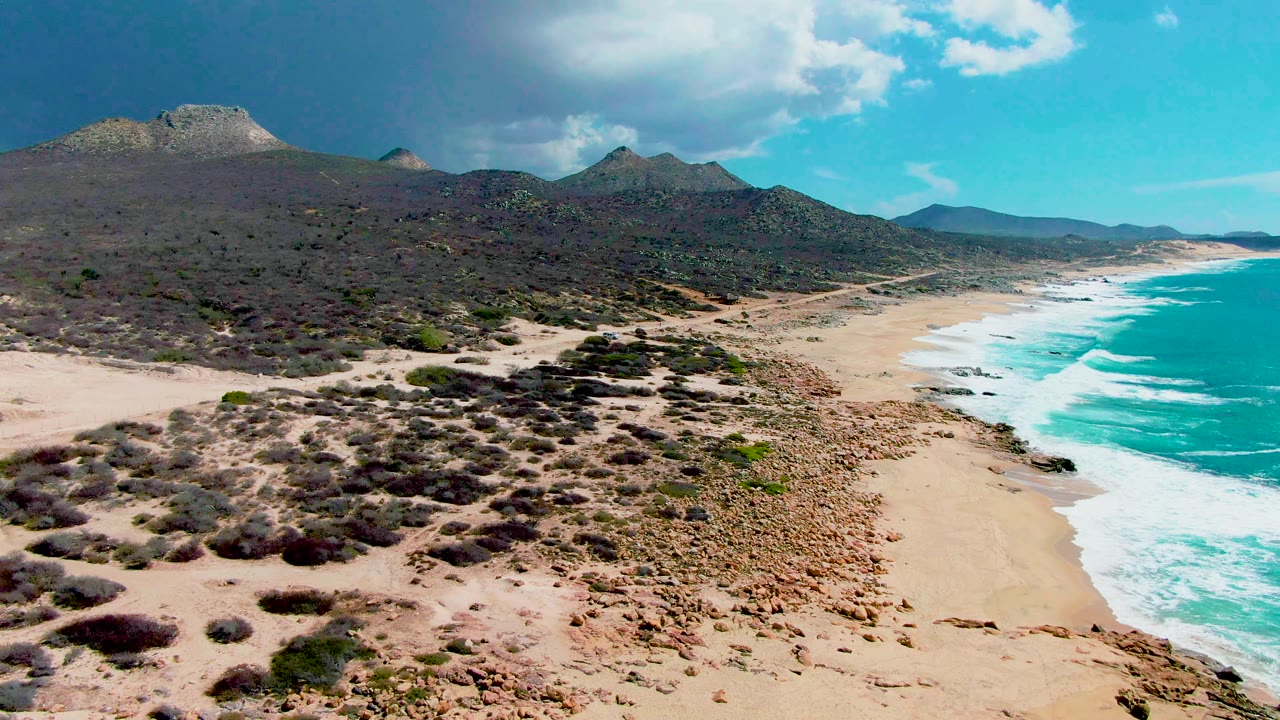 Wide aerial shoreline with desert scrub, mountains, and white waves.