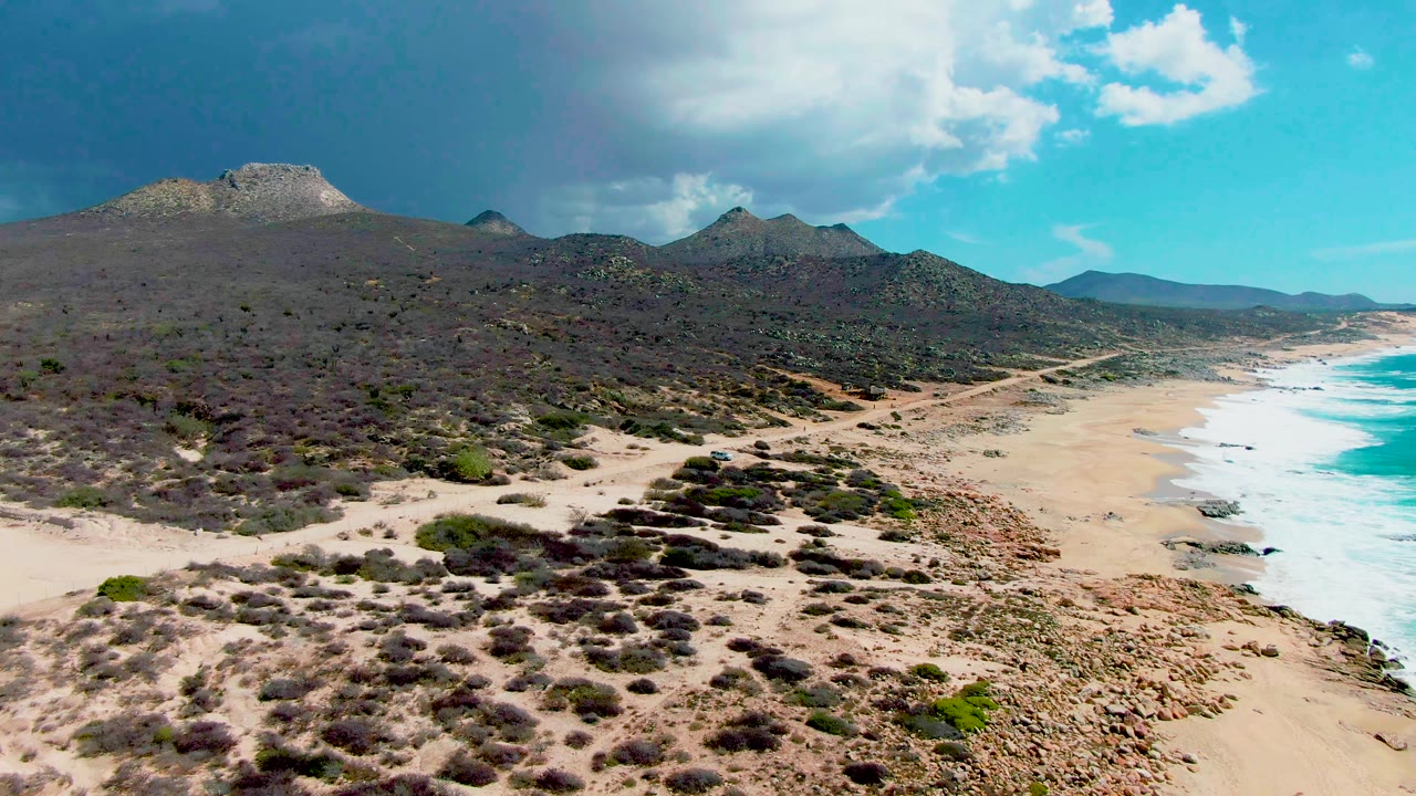 Aerial view at Punta Buzzards, Baja California Sur, showing a dirt road running beside a sandy beach, turquoise surf, scrubland, mountains, and no visible people.