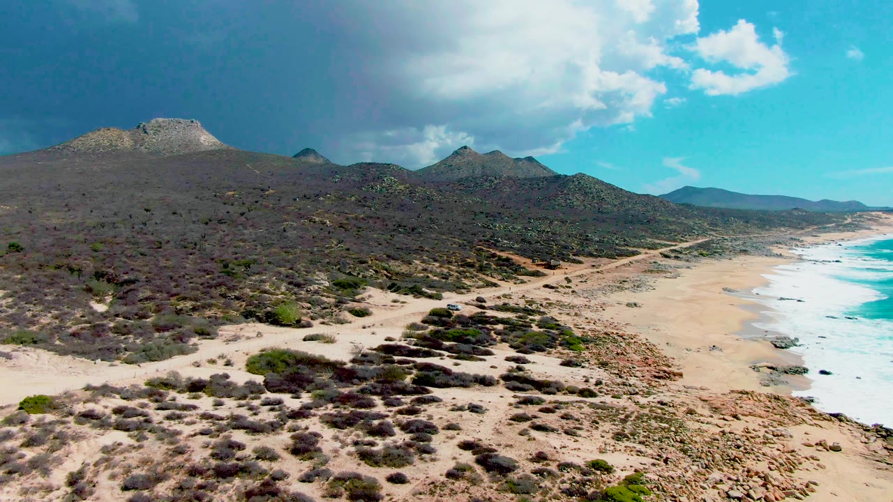 Aerial frame at Punta Buzzards with the dirt road more centered through scrubland, a small vehicle near the lower road, sandy beach to the right, and no visible people.