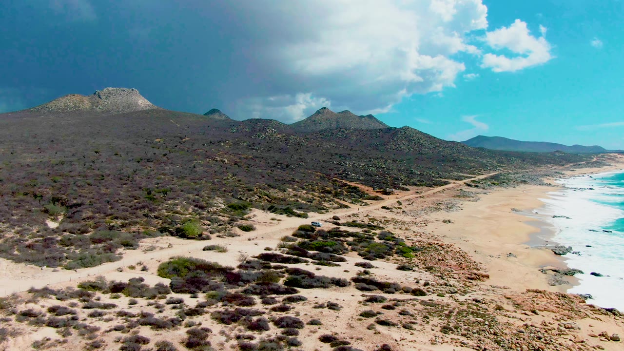 Punta Buzzards aerial view with the small vehicle visible on the lower dirt road, scrub-covered hills filling the left side, surf on the right, and no visible people.