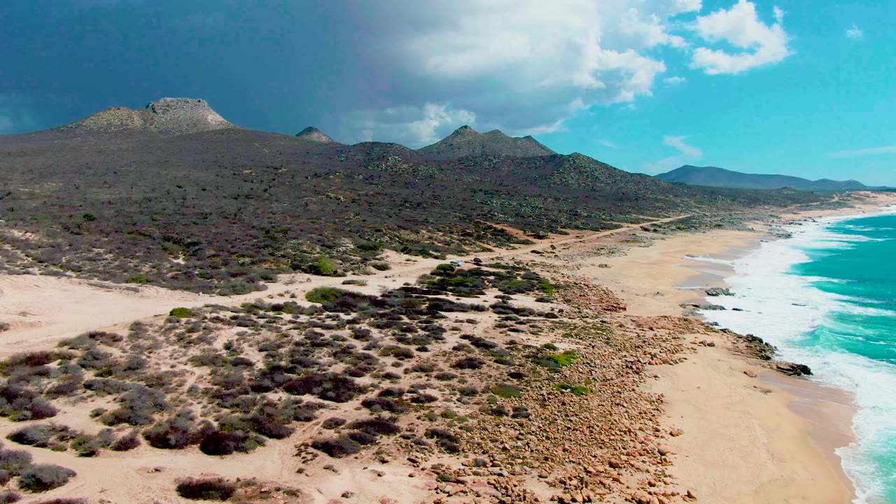 Aerial frame over Punta Buzzards showing sandy beach and turquoise surf on the right, scrubby desert terrain in the foreground, low mountains in back, and no visible people.