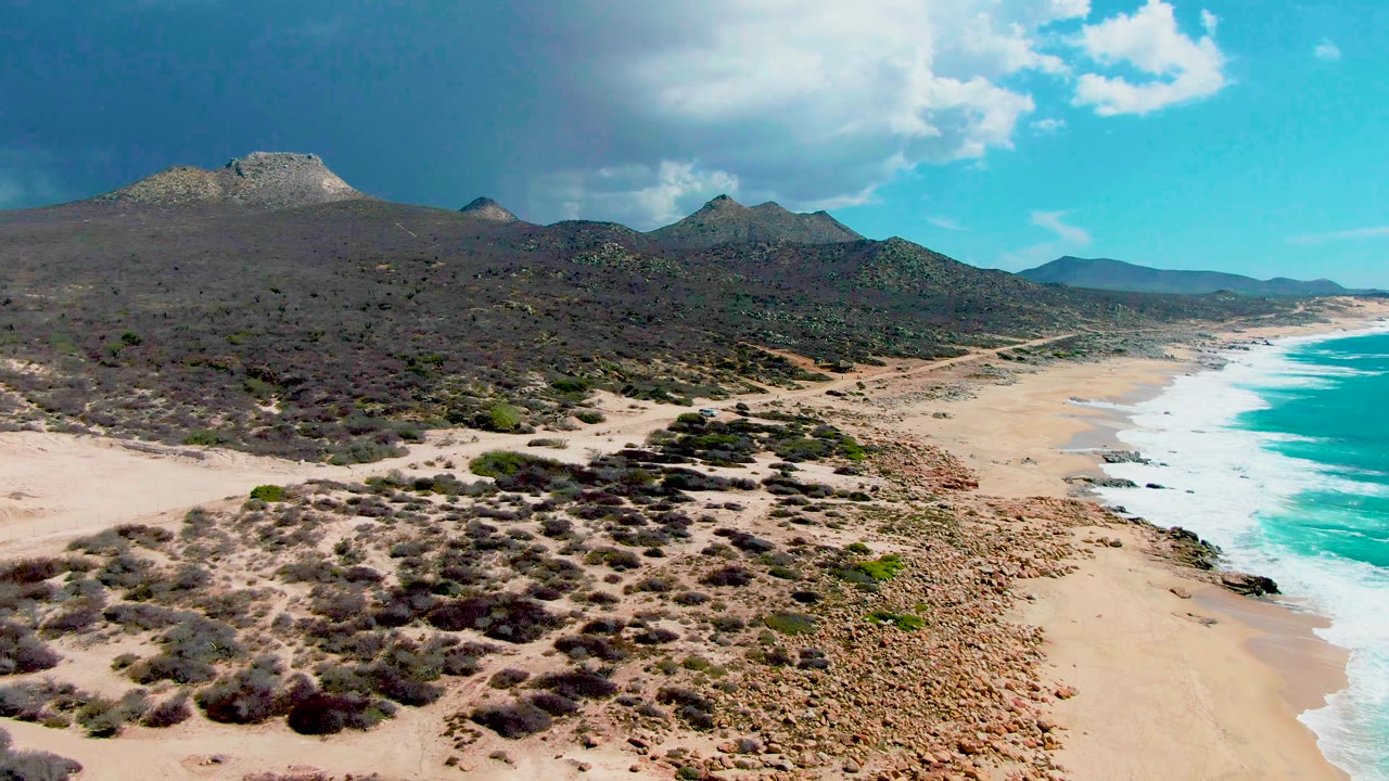 Aerial view at Punta Buzzards with scrub-covered dunes filling more of the foreground, the sandy beach curving beside turquoise waves, distant hills, and no visible people.