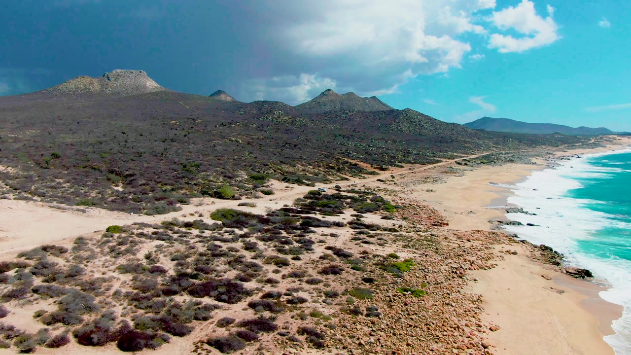 Aerial frame over Punta Buzzards with the beach and white surf pushed to the right edge, dry shrubland across the center, mountains on the horizon, and no visible people.