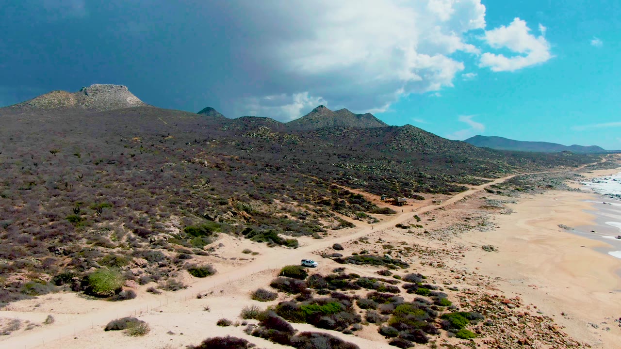 Aerial frame at Punta Buzzards, Baja California Sur, showing a sandy dirt road crossing desert scrub beside a broad beach, with low mountains and no visible people.