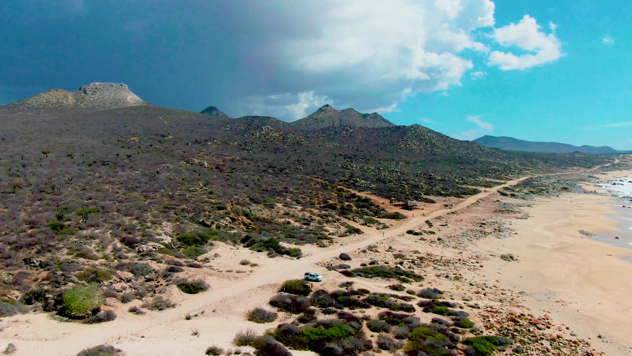 Wide aerial view in Punta Buzzards with the light-colored car lower in frame on the sandy track, beach to the right, desert hills ahead, and no visible people.