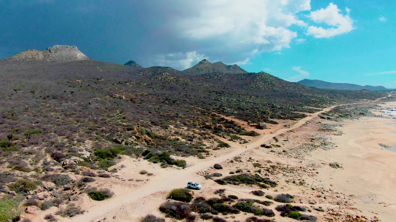 Drone view of desert hills and a beach road under bright clouds.