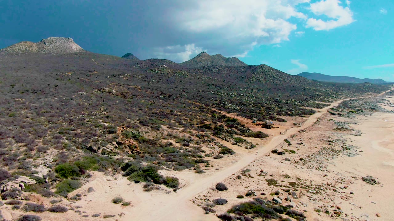 Aerial frame from Punta Buzzards, Baja California Sur showing a pale dirt road crossing arid shrubland toward distant mountains under a partly cloudy blue sky.