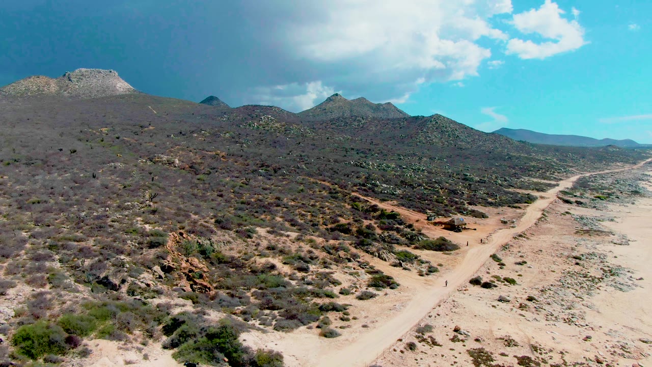 Aerial frame over Punta Buzzards desert terrain where the pale road angles along the right side, with shrubs, rocky patches, and mountain ridges in the background.