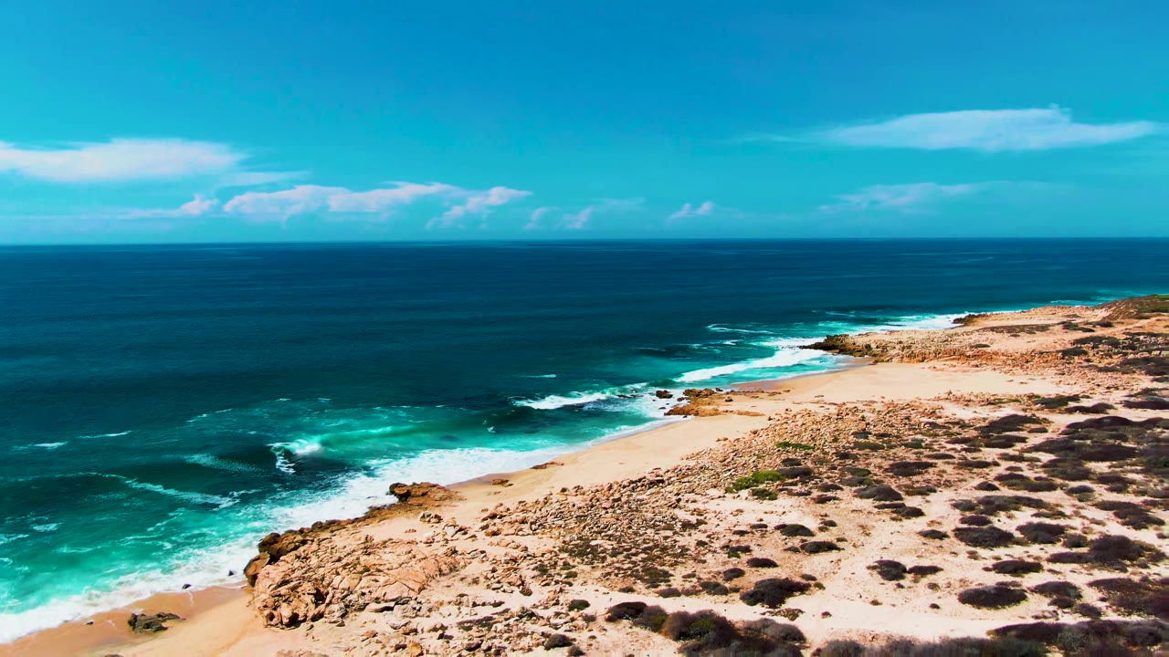 Aerial view of Punta Buzzards coastline with turquoise surf breaking beside rocky sand and low scrub, with no visible people in this opening frame.