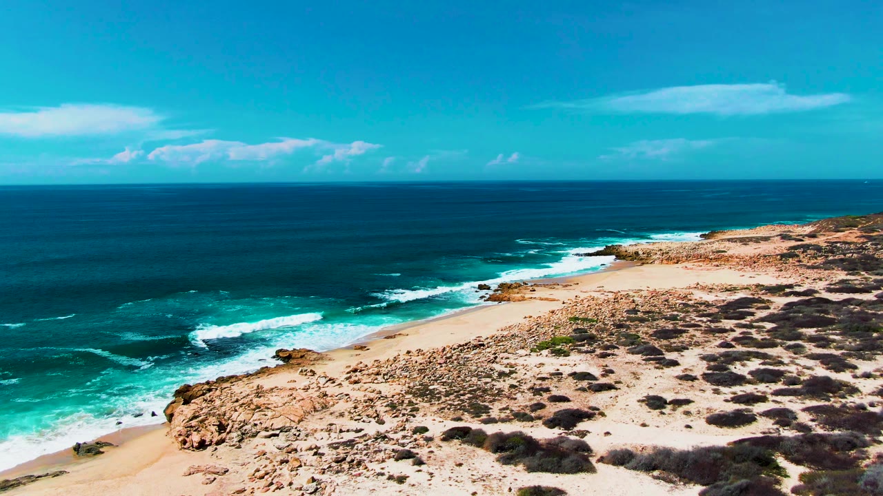 Drone-height view of Punta Buzzards with darker blue offshore water, turquoise nearshore bands, white foam, and a rocky sandy beach stretching to the right, with no visible people.