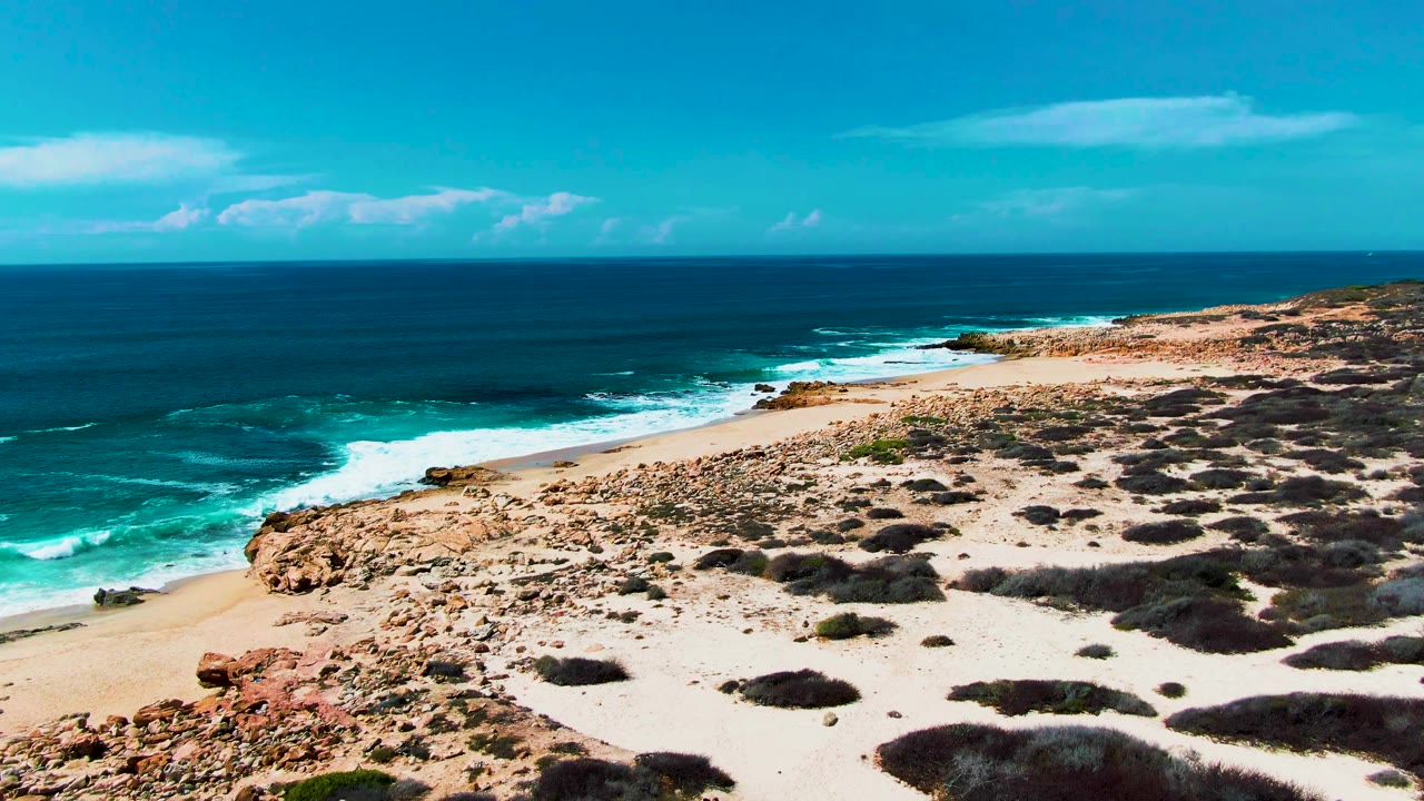 At 0:00, Punta Buzzards shoreline in Baja California Sur shows turquoise surf breaking beside rocks, pale sand, low scrub, and no visible people.