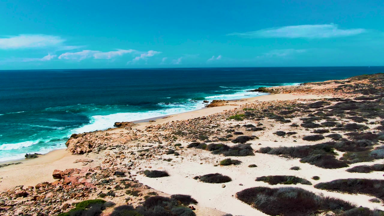 At 0:01, the elevated view holds on the Punta Buzzards coast as white water spreads along the beach edge with no visible people.