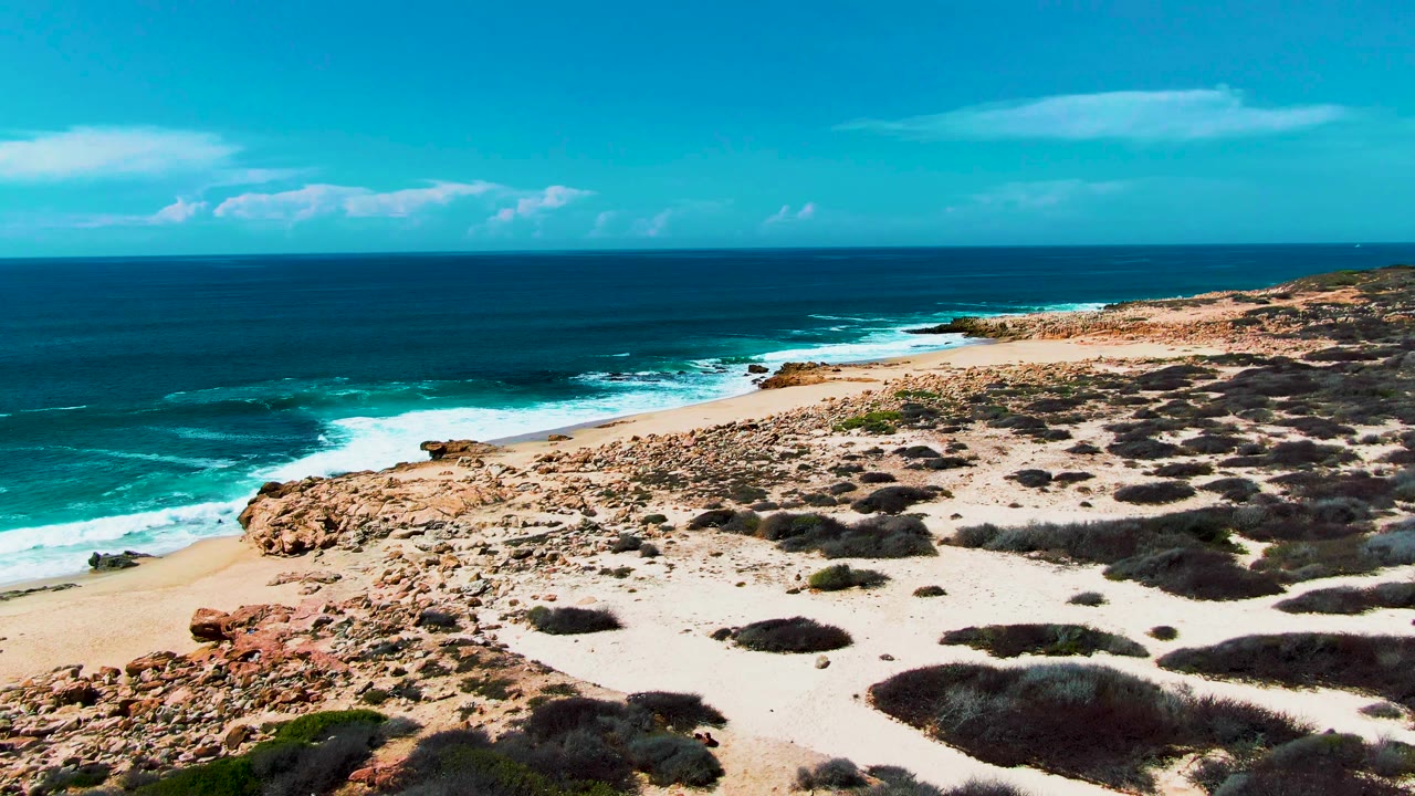 At 0:02, the Baja California Sur shoreline remains clear and empty, with rocky sand in the foreground and turquoise surf below the horizon.