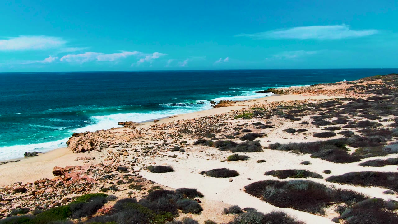 Wide aerial coastal view with dark blue sea, white foam, sandy beach, rocks, and scrub.