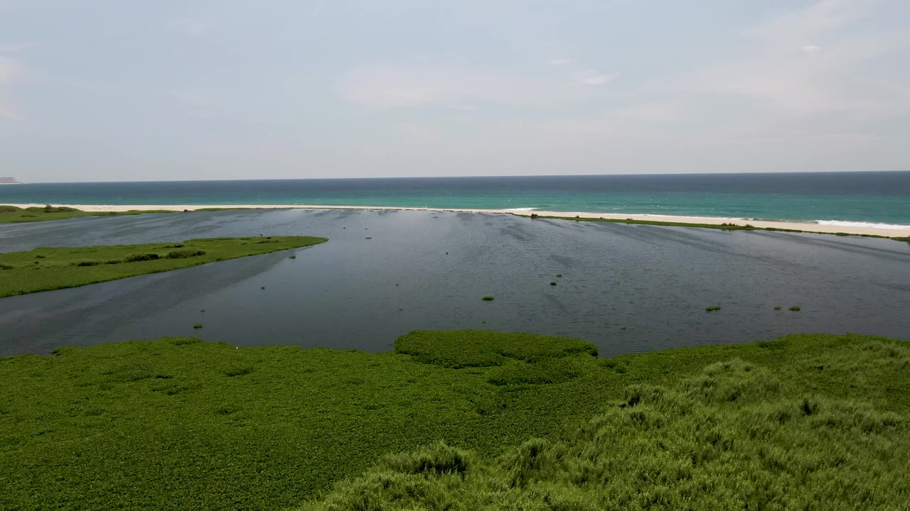 At two seconds, the San Jose Del Cabo tidal flat sits under soft daylight, with darker shallow water, green vegetation patches, a pale beach strip, turquoise surf, and no visible people.