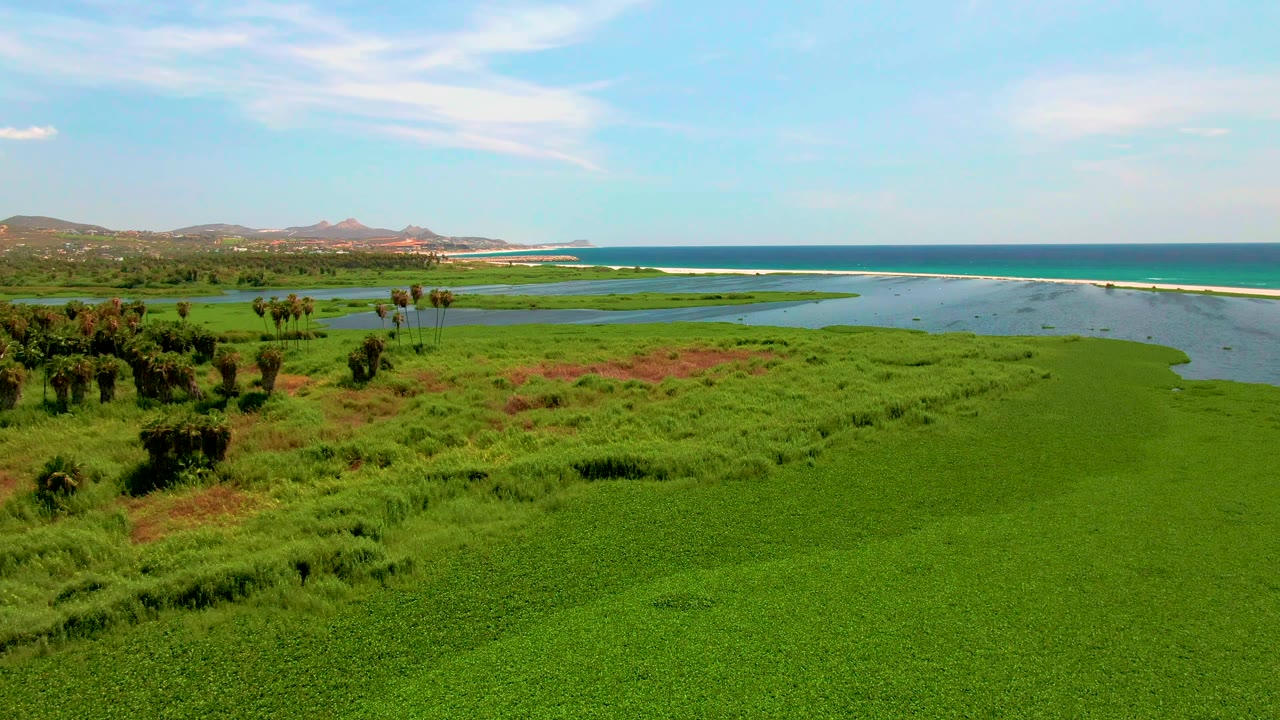 At 0:02, the San Jose Del Cabo wetland spreads across the foreground with a curving water channel, a narrow white surf line, blue ocean, distant hills, light clouds, and no visible people.