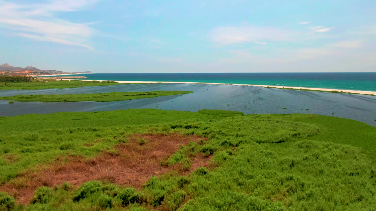 At 0:02, the San Jose del Cabo lagoon spreads across the center with thick green vegetation below, a pale sandbar and surf line beyond it, low hills at left, and no visible people.