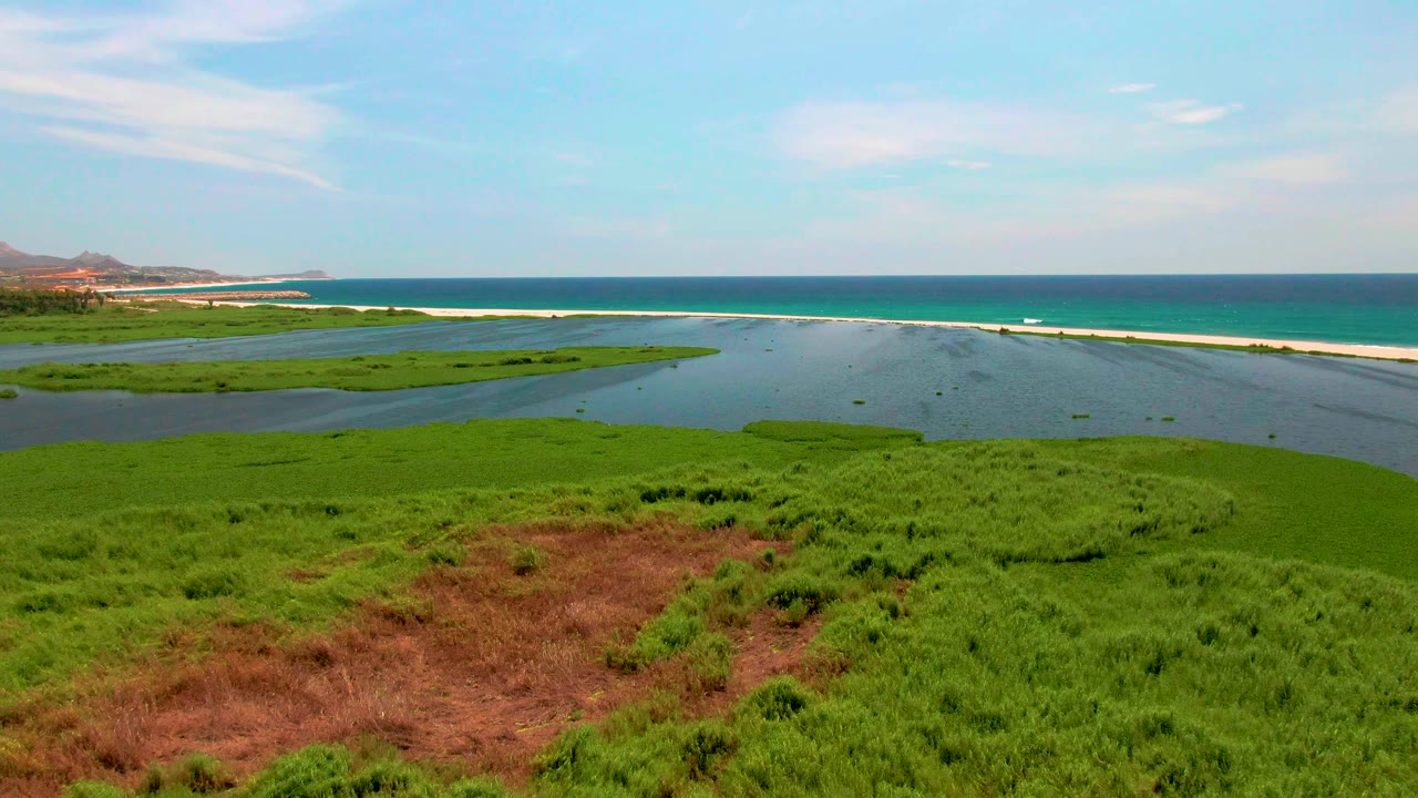 Midway through the clip, the San Jose del Cabo wetland fills the foreground with a larger brown patch, while the lagoon, sandbar, ocean horizon, distant hills, and scattered clouds remain visible with no people.