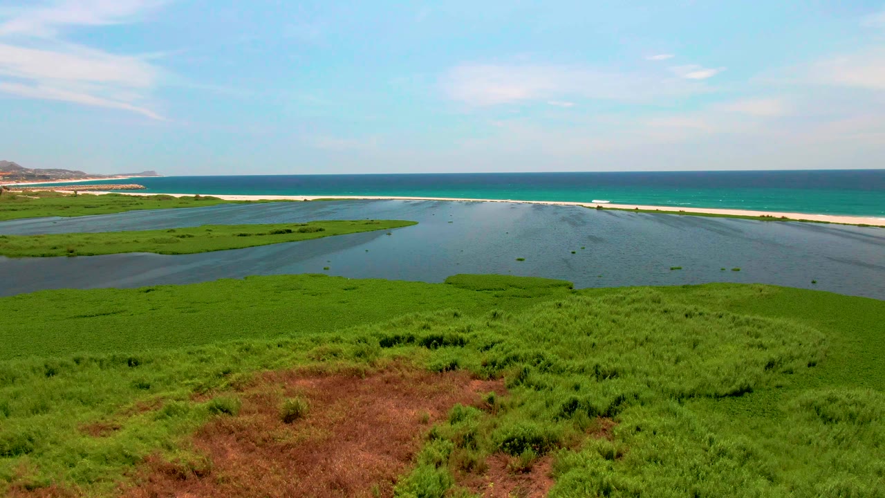 Aerial frame from San Jose del Cabo, Mexico, showing green marsh in the foreground, lagoon water beyond it, a pale sandbar, blue ocean, and no visible people.