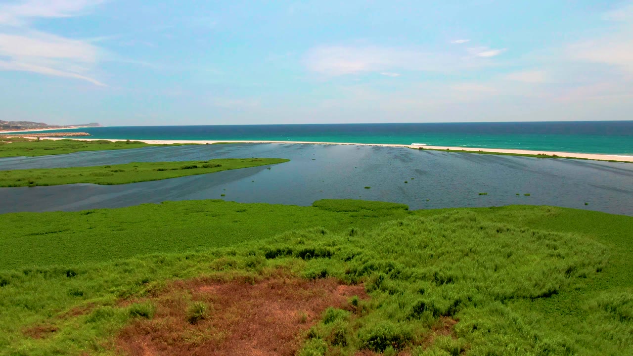 Aerial view in San Jose del Cabo, Mexico, with dense green marsh grass below, calm lagoon water in the center, white surf along the sandbar, and no visible people.
