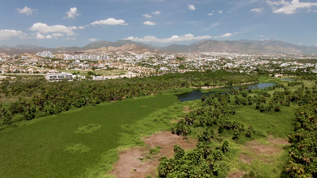 Wide aerial landscape with green field, pond, buildings, and mountains