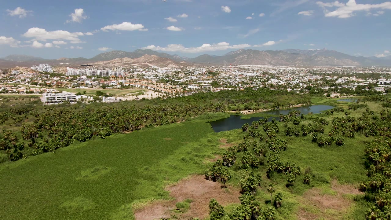 At two seconds, the aerial view keeps the tree grove and ponds centered below the San Jose Del Cabo town edge, with dry mountains and a partly cloudy sky in the background and no visible people.