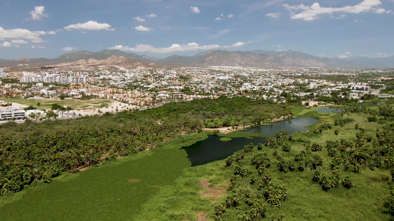 Opening aerial frame over San Jose del Cabo with green fields and tree cover in the foreground, small ponds near the city edge, distant mountains, and no visible people.