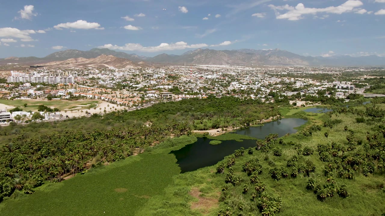 Wide drone view of a greenbelt, dark pond, white city buildings, and mountains.