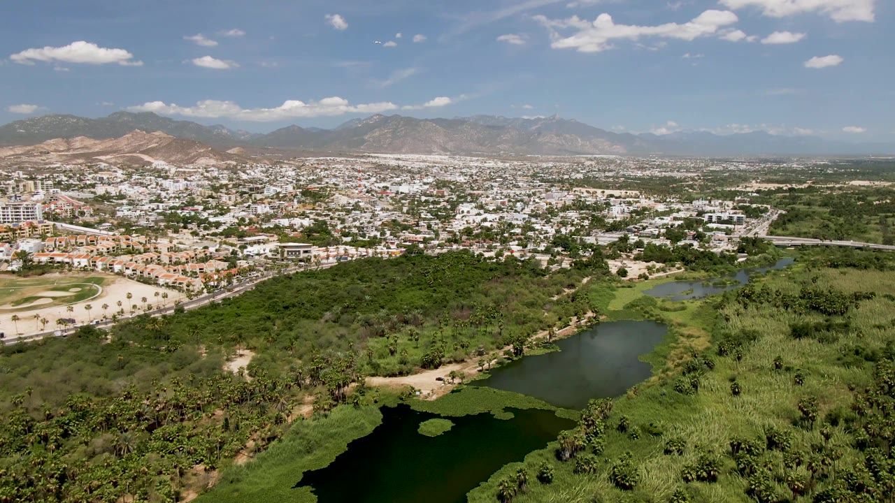 Wide aerial view over San Jose del Cabo with a dark pond and green parkland in the foreground, city buildings beyond, mountains on the horizon, and no visible people.