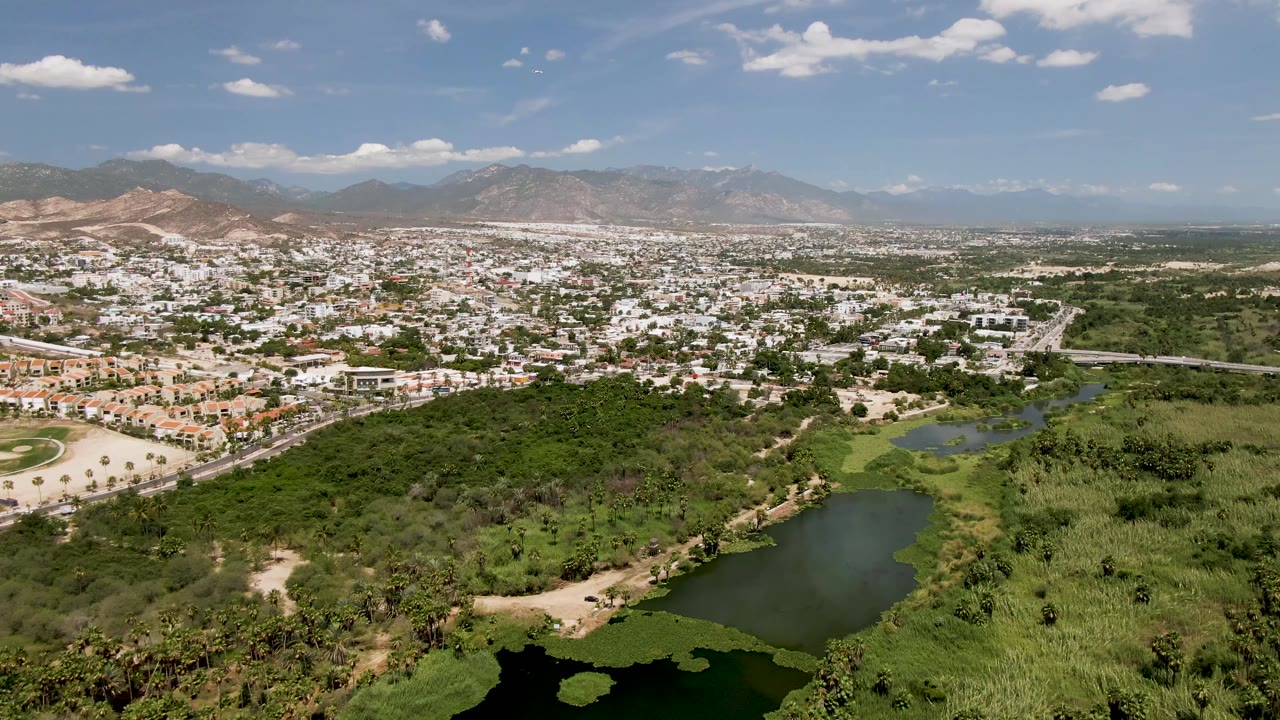 High-angle view of San Jose del Cabo with a pond low in frame, thick green vegetation, light-colored neighborhoods, dry hills and mountains behind, and no visible people.