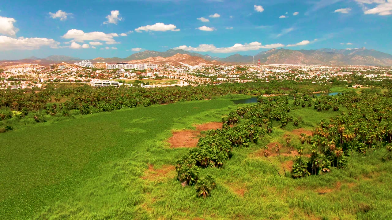 Early pan frame in San Jose del Cabo showing a broad green field, dense tree clusters, red soil, distant buildings, and hazy mountains under scattered clouds, no visible people.