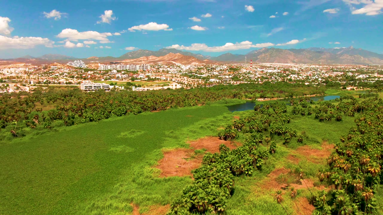 Later aerial frame in San Jose del Cabo where green fields lead toward tree cover, a small blue water area, distant buildings, and mountain slopes, no visible people.