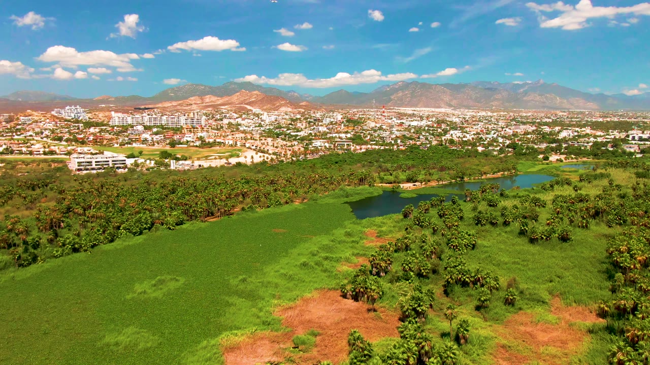 Opening aerial frame over San Jose Del Cabo with broad green fields in front, a small water body near the town, distant mountains, scattered clouds, and no visible people.