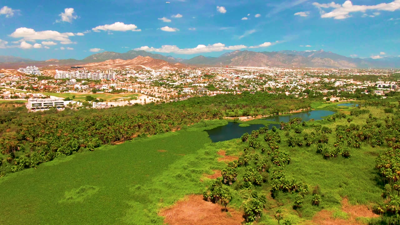 Aerial view across green terrain toward white buildings and distant mountains.